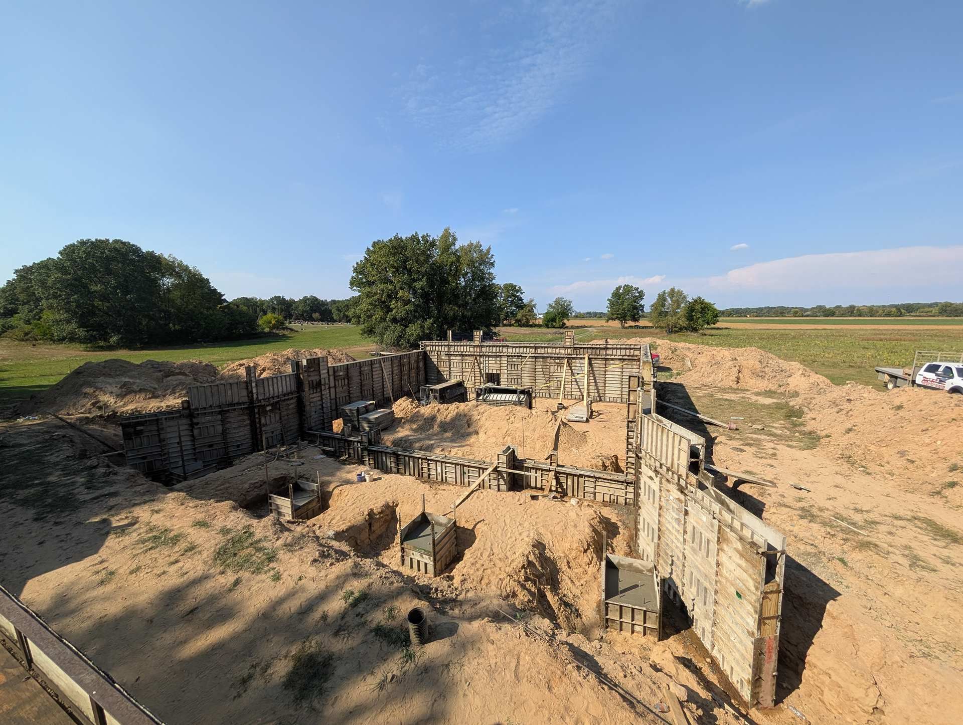 Concrete foundation walls and footings for a building under construction in a sandy, open field under a blue sky.