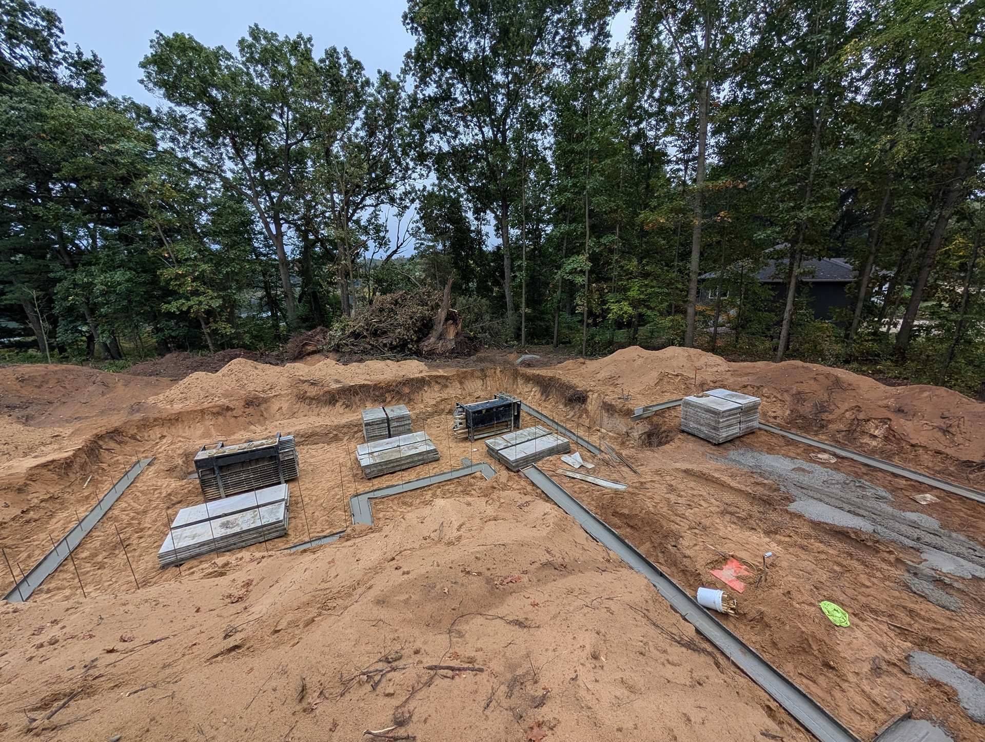 A construction site with concrete foundation forms and stacks of cinder blocks on sandy ground, backed by dense trees.