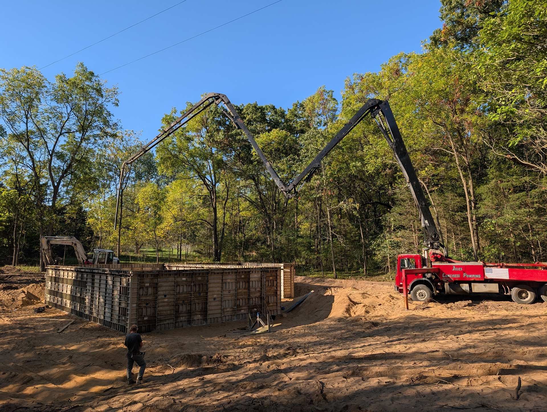 A red concrete pump truck extends its boom over wooden foundation forms at a construction site in a wooded area.