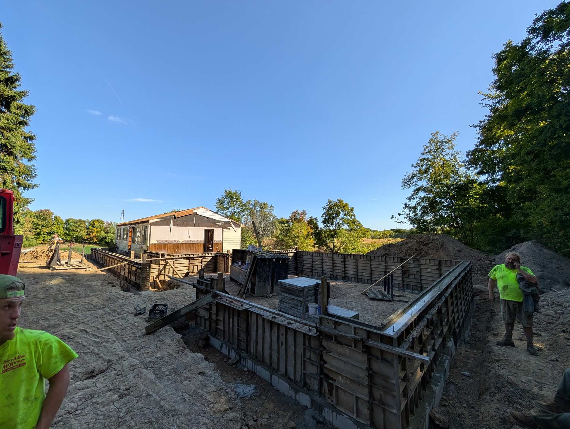 Two workers in high-visibility neon shirts stand at a construction site with concrete foundation forms for a new house.