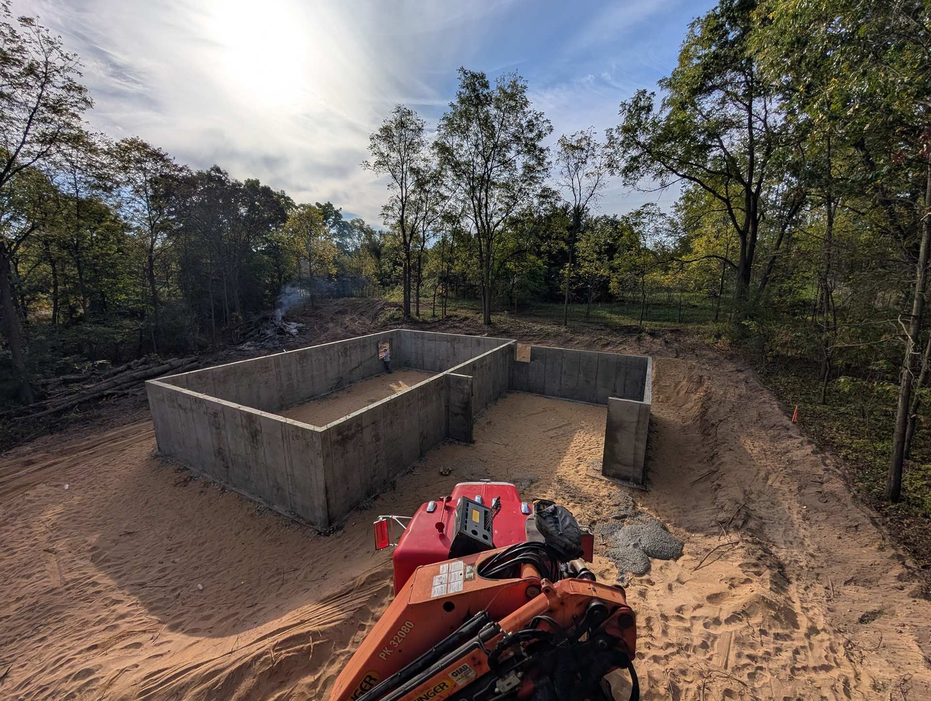 A foundation of grey concrete walls sits on a sandy, cleared construction site surrounded by trees on a sunny day.