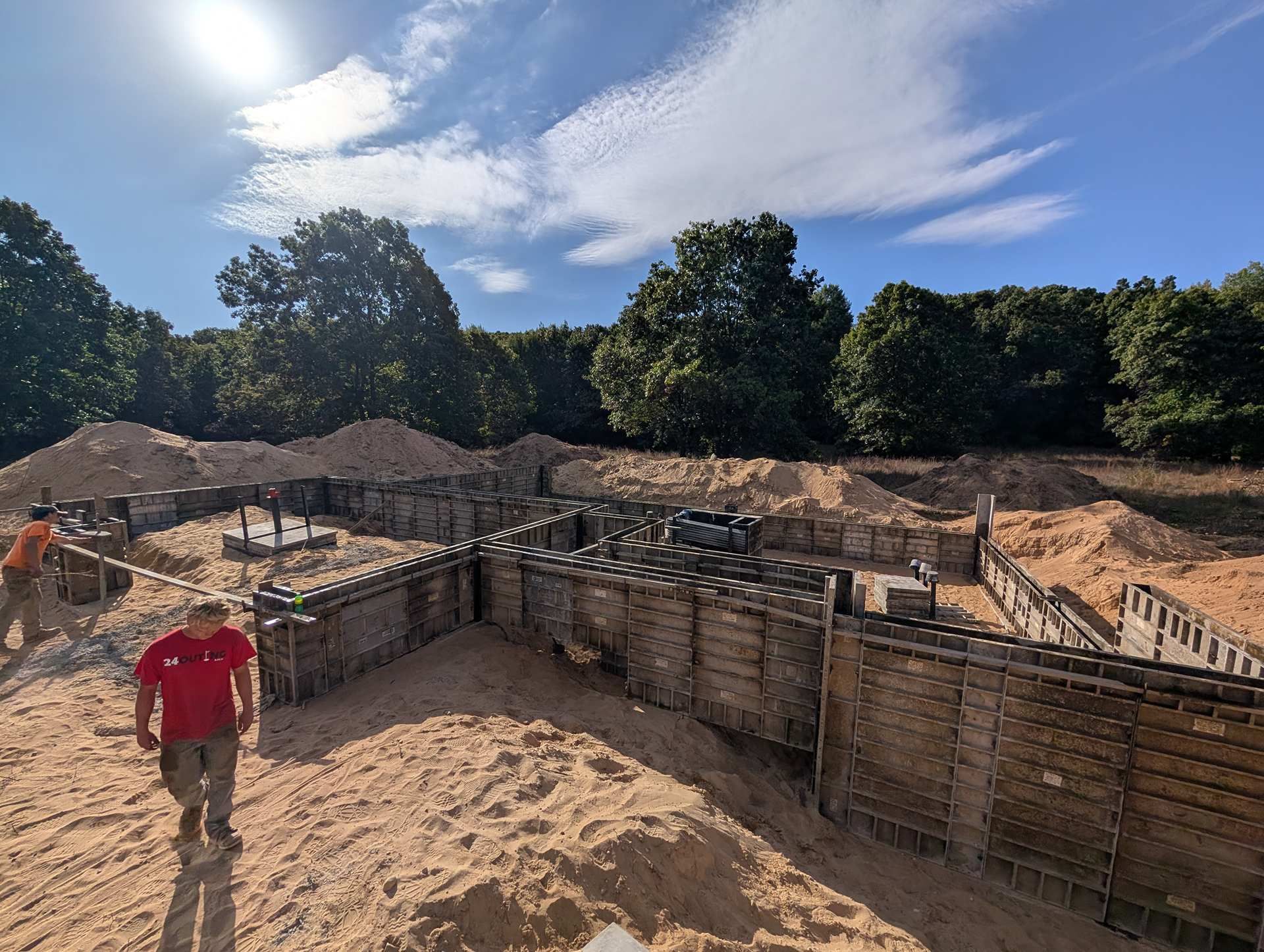 A construction worker in a red shirt walks past concrete formwork set up for a building foundation on a sandy site.