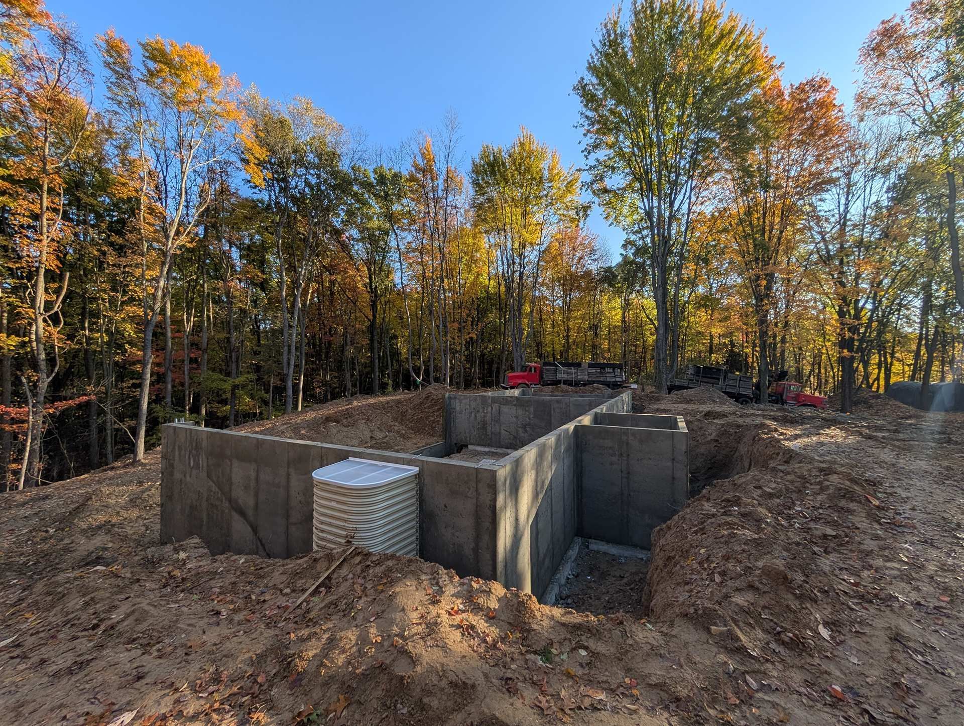 A concrete foundation for a new house under construction, surrounded by a forest with autumn foliage under a blue sky.
