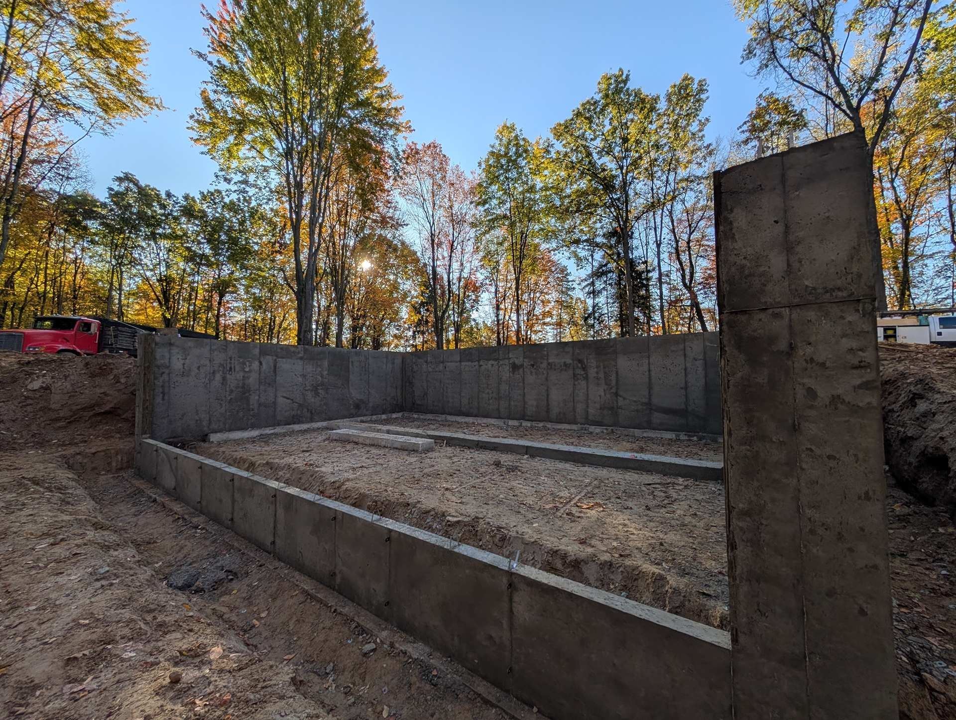 A newly poured concrete foundation for a building site surrounded by autumn trees under a clear blue sky.