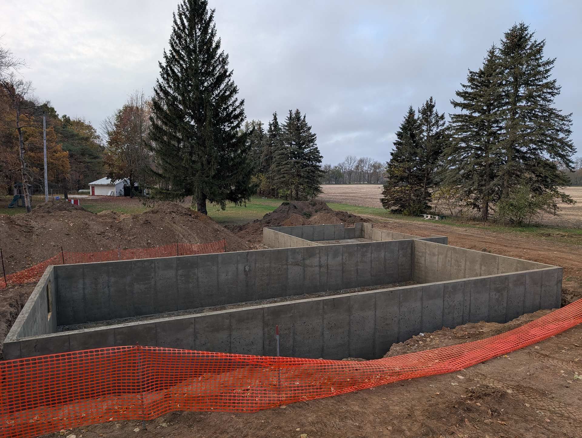 A concrete foundation for a new building sits in an open field, surrounded by orange safety fencing and pine trees.