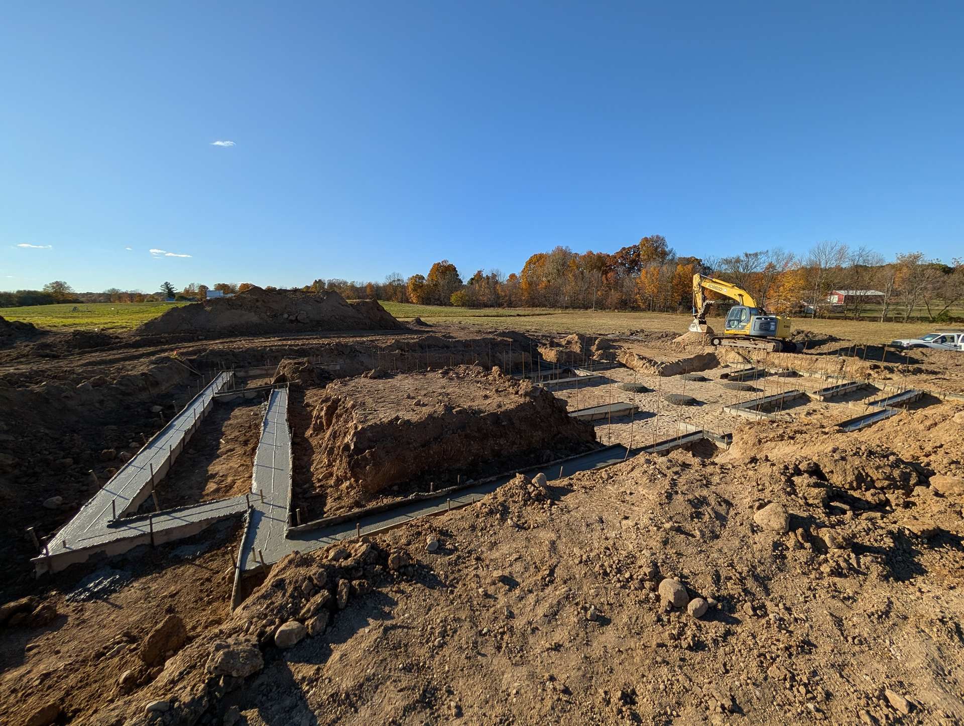 A construction site showing concrete foundation footings in a dirt field with an excavator in the distance.