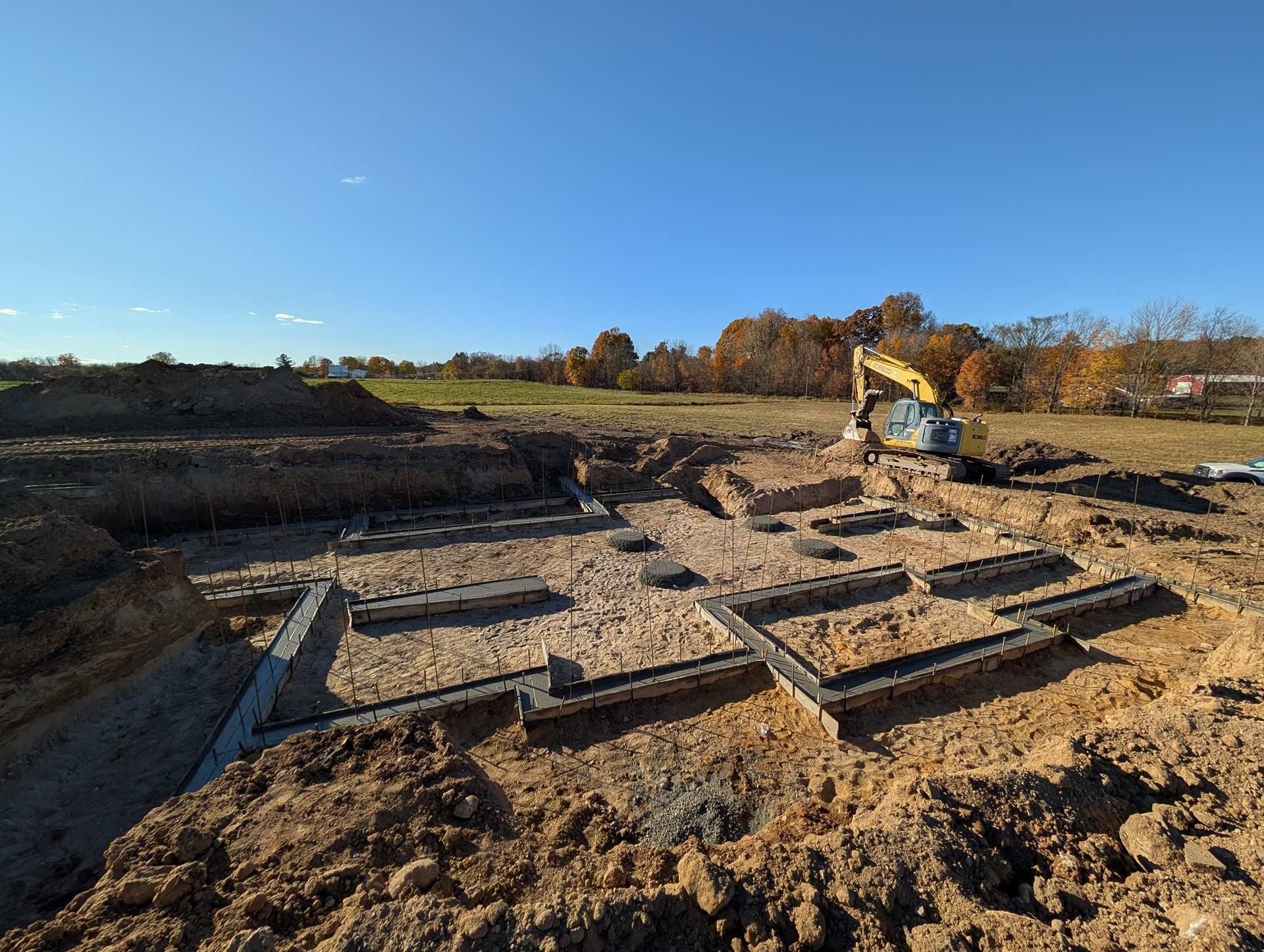 A yellow excavator works at a construction site featuring concrete foundation footings set in a dirt lot under blue skies.