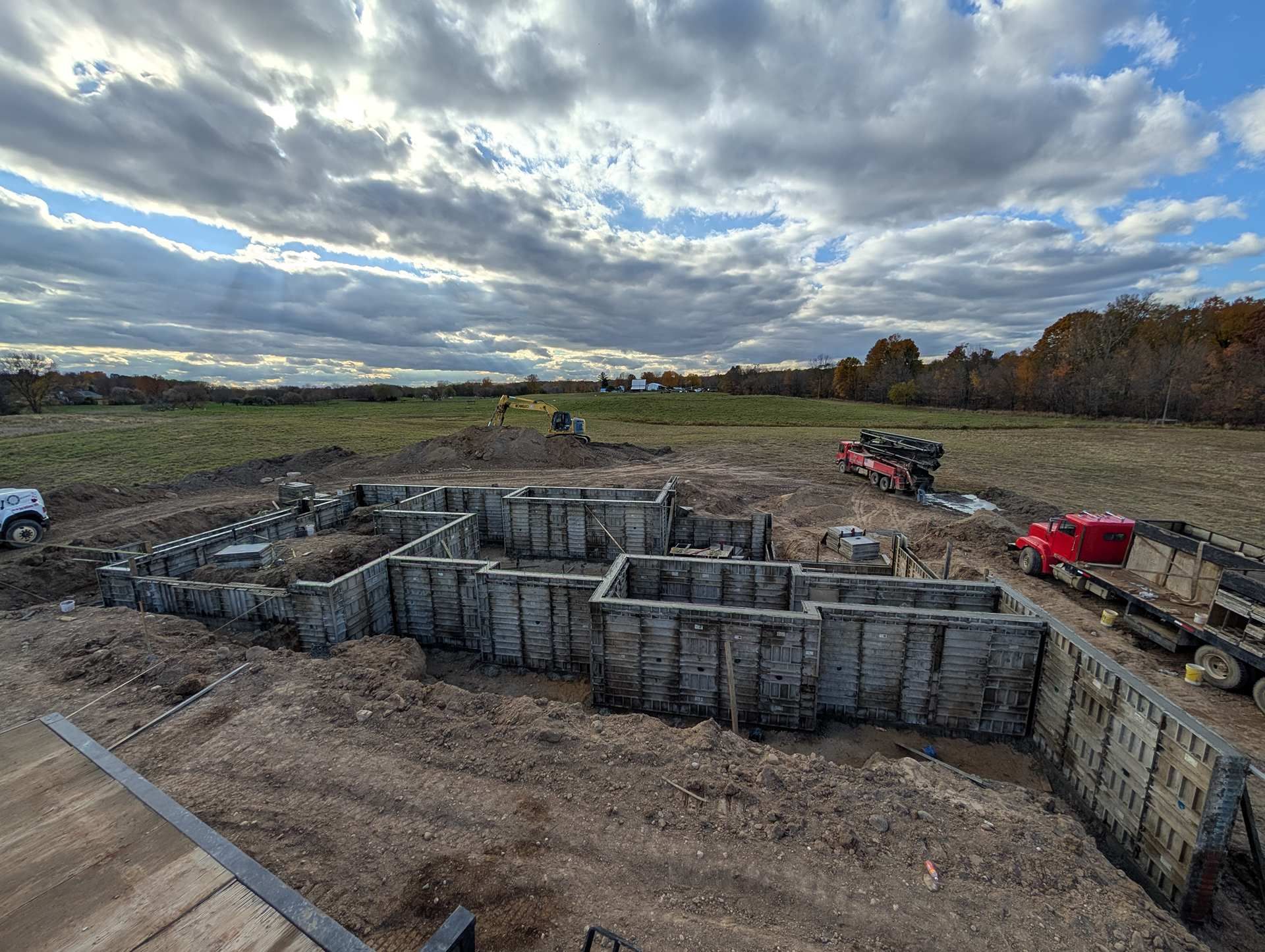 A construction site showing concrete foundation forms installed for a building, with equipment nearby under a cloudy sky.