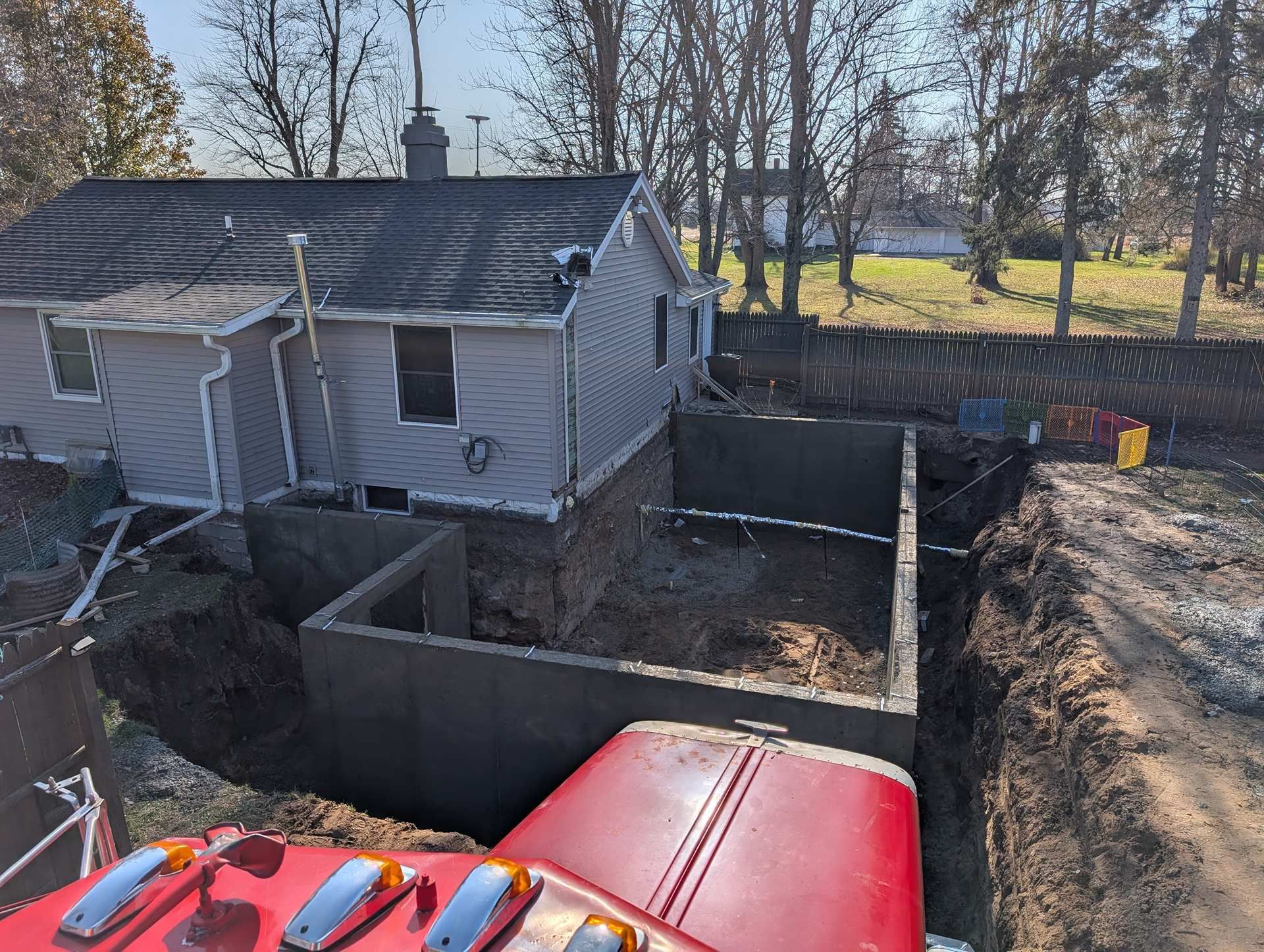 A view from a construction vehicle shows a house foundation addition being built, with gray walls set into a dirt trench.