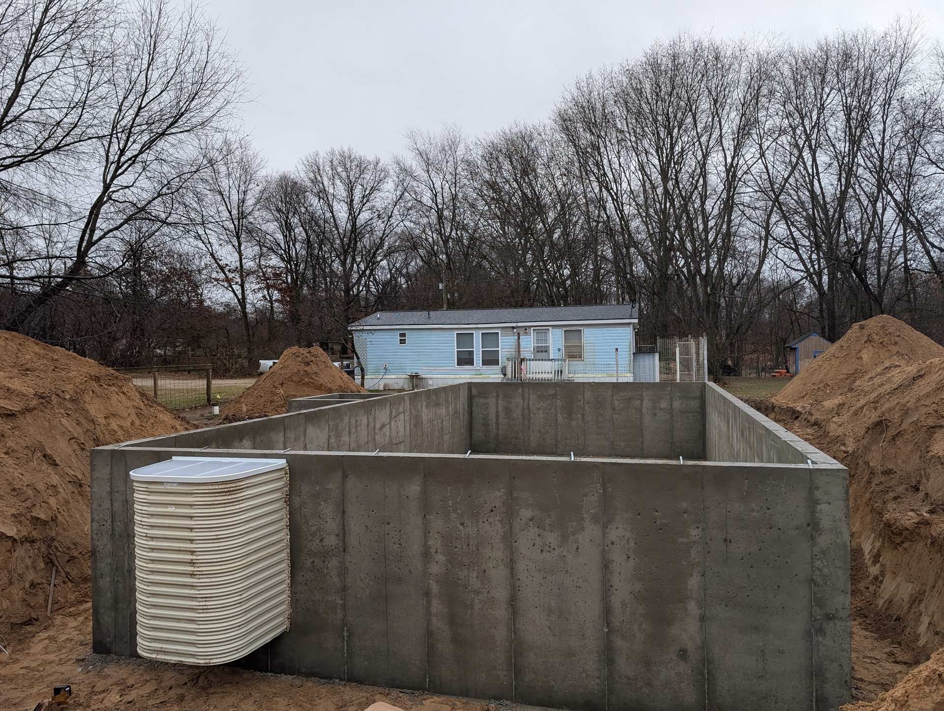 A freshly poured concrete foundation for a house, with a window well attached to the exterior wall, under a cloudy sky.