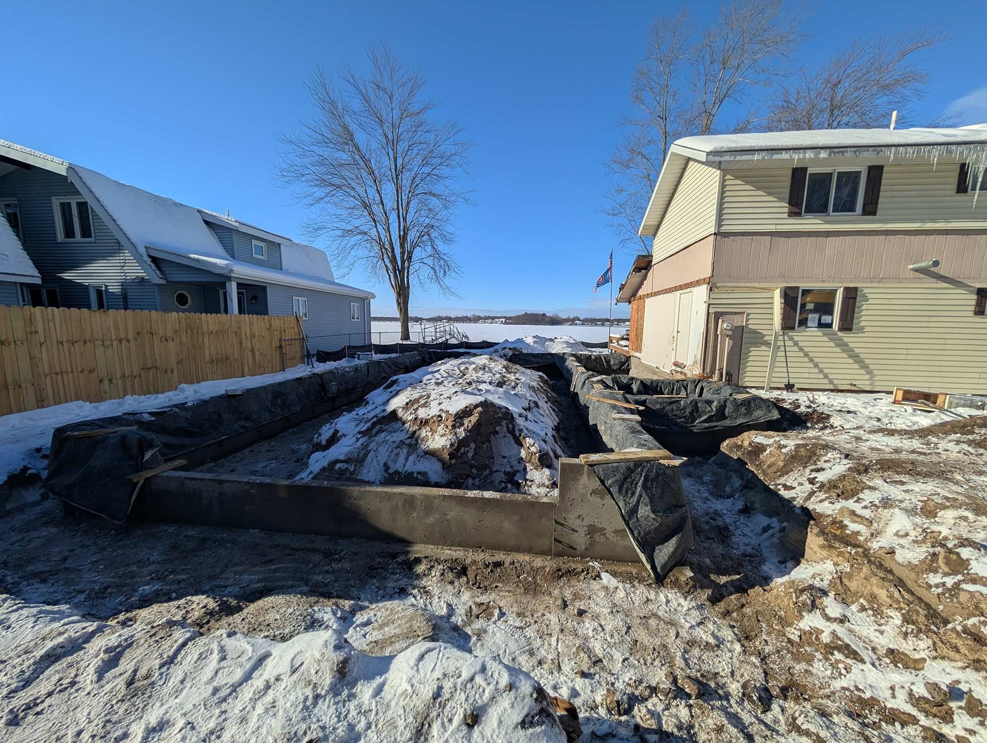 A concrete foundation poured in a snowy yard between two houses under a clear blue sky.