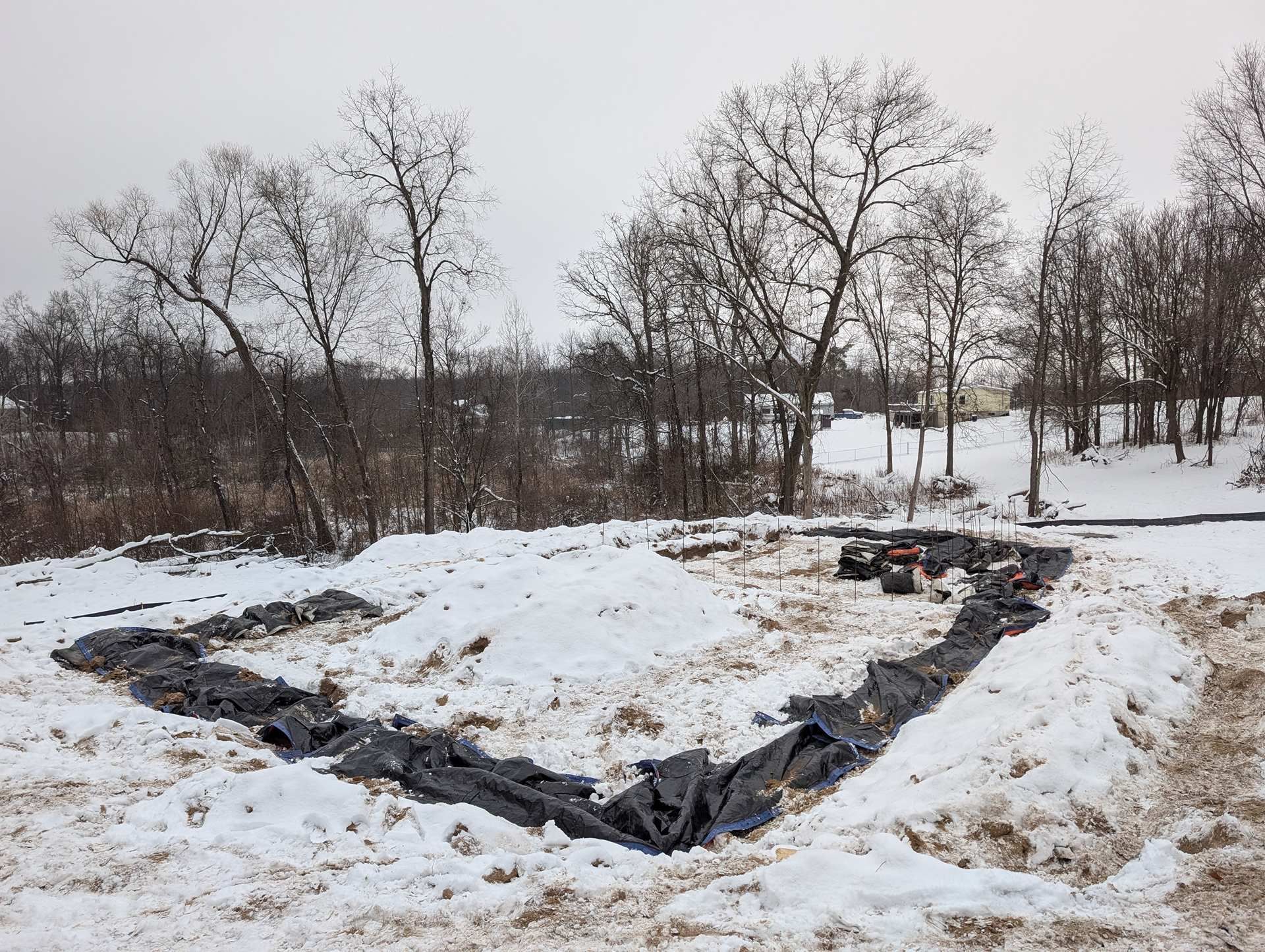 A construction site in the snow featuring a rectangular trench lined with black plastic sheeting.
