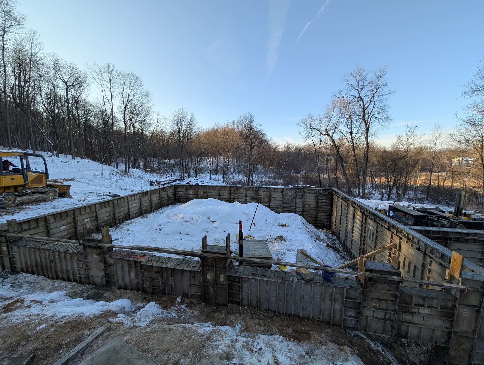 A concrete foundation for a building under construction, surrounded by snow and trees on a sunny winter day.