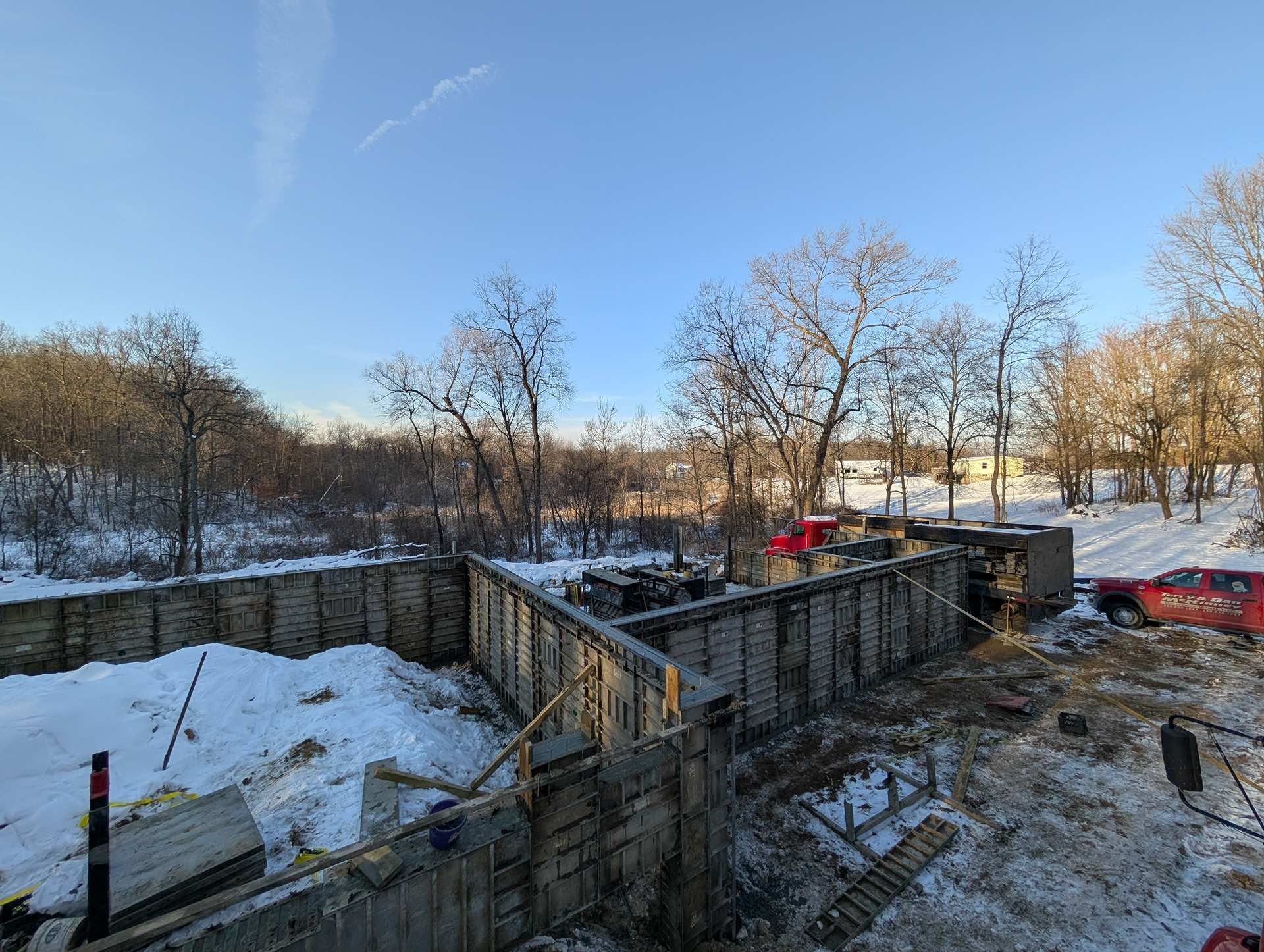 A red pickup truck parked at a construction site with concrete foundation walls surrounded by snow and bare trees.