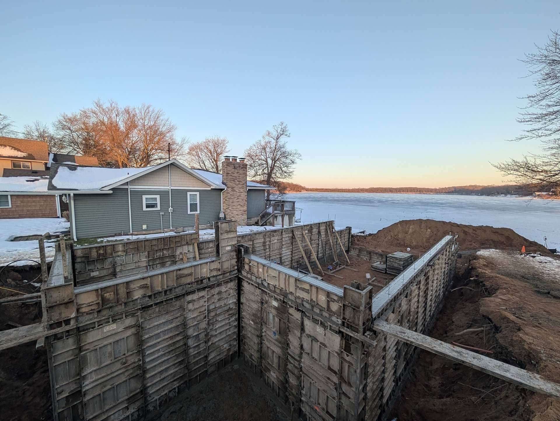 Concrete foundation forms for a building under construction, set on a snowy lot next to a house and a frozen lake.