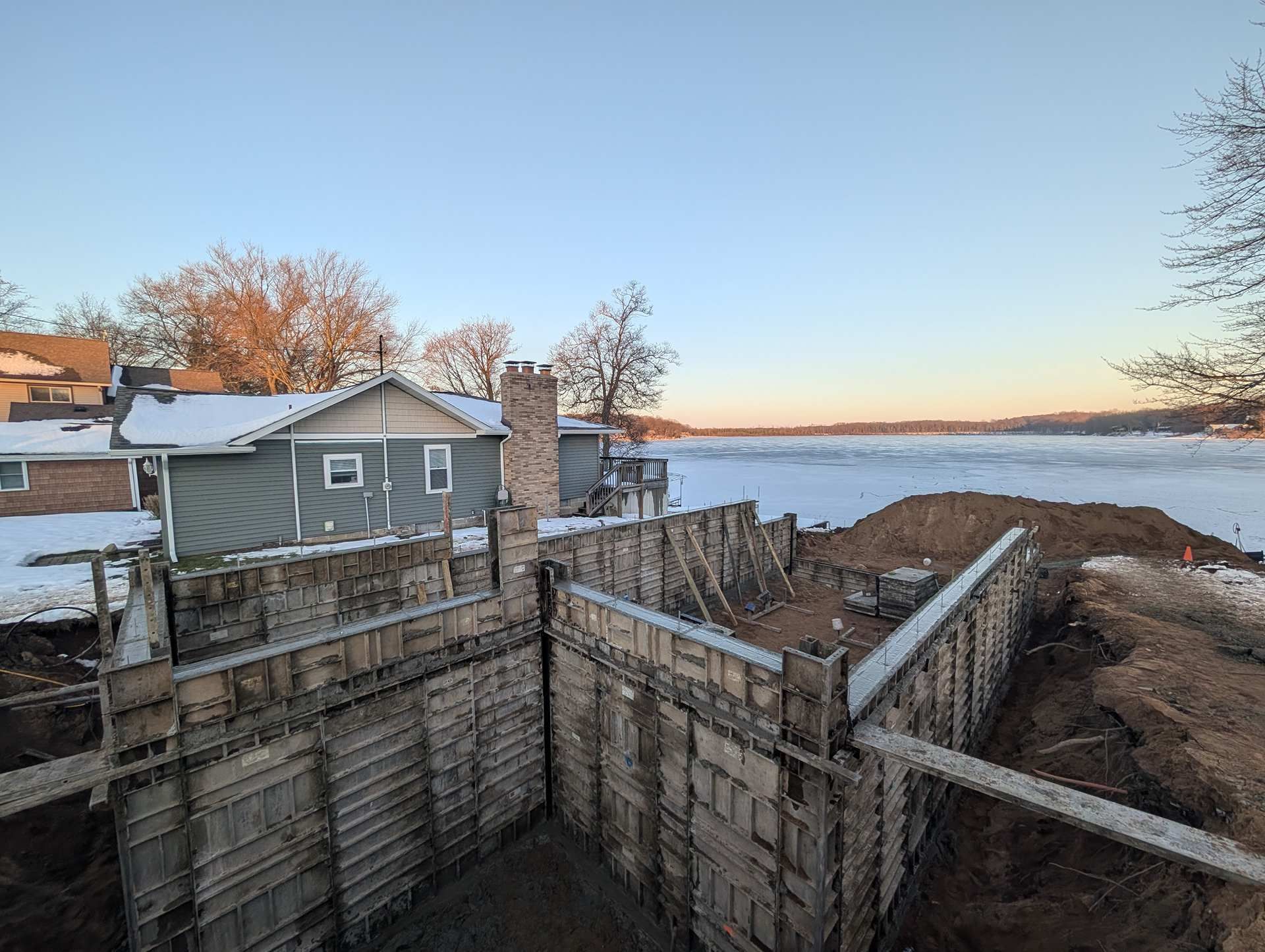 Concrete foundation forms for a home addition, viewed from an excavation site next to a snowy house and frozen lake.