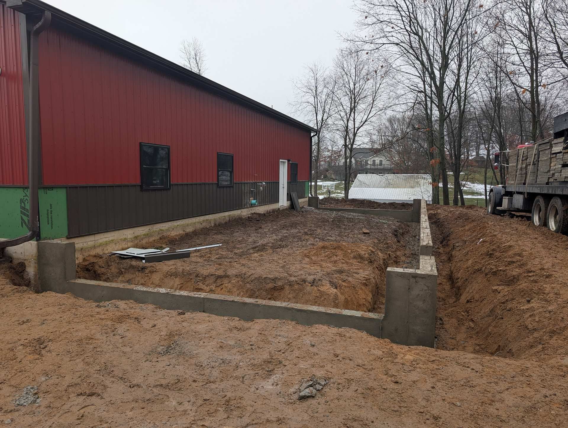 A partially constructed concrete foundation for an building extension, set in a dirt lot beside a large red metal barn.