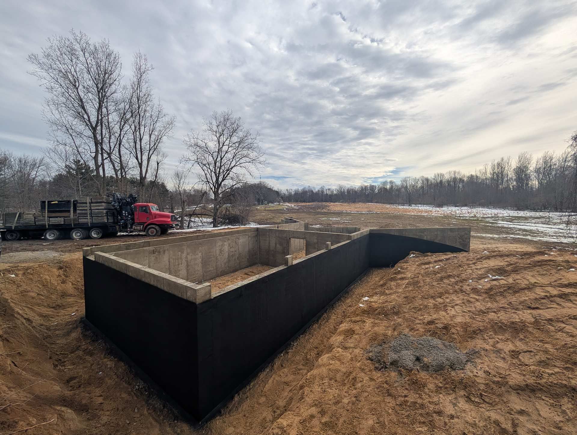 A red truck sits near a concrete foundation wall with a black waterproofing coating on a construction site.