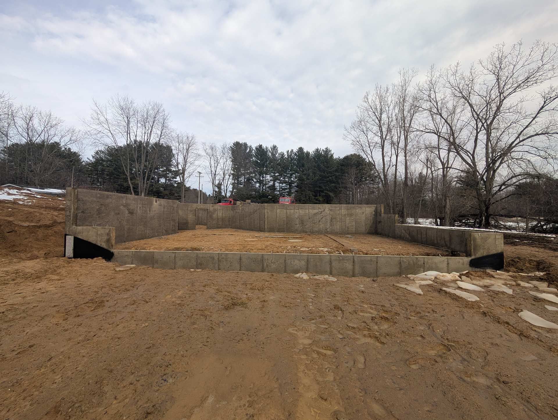 Concrete foundation walls for a building under construction on a dirt lot with bare trees in the background.