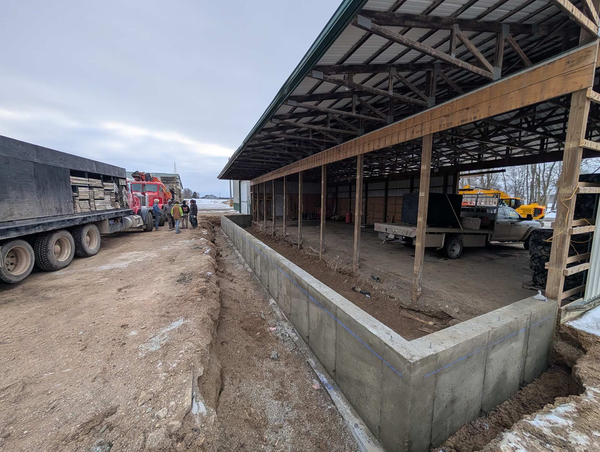 A large shed with a concrete foundation under construction next to a flatbed truck on a gravel lot.