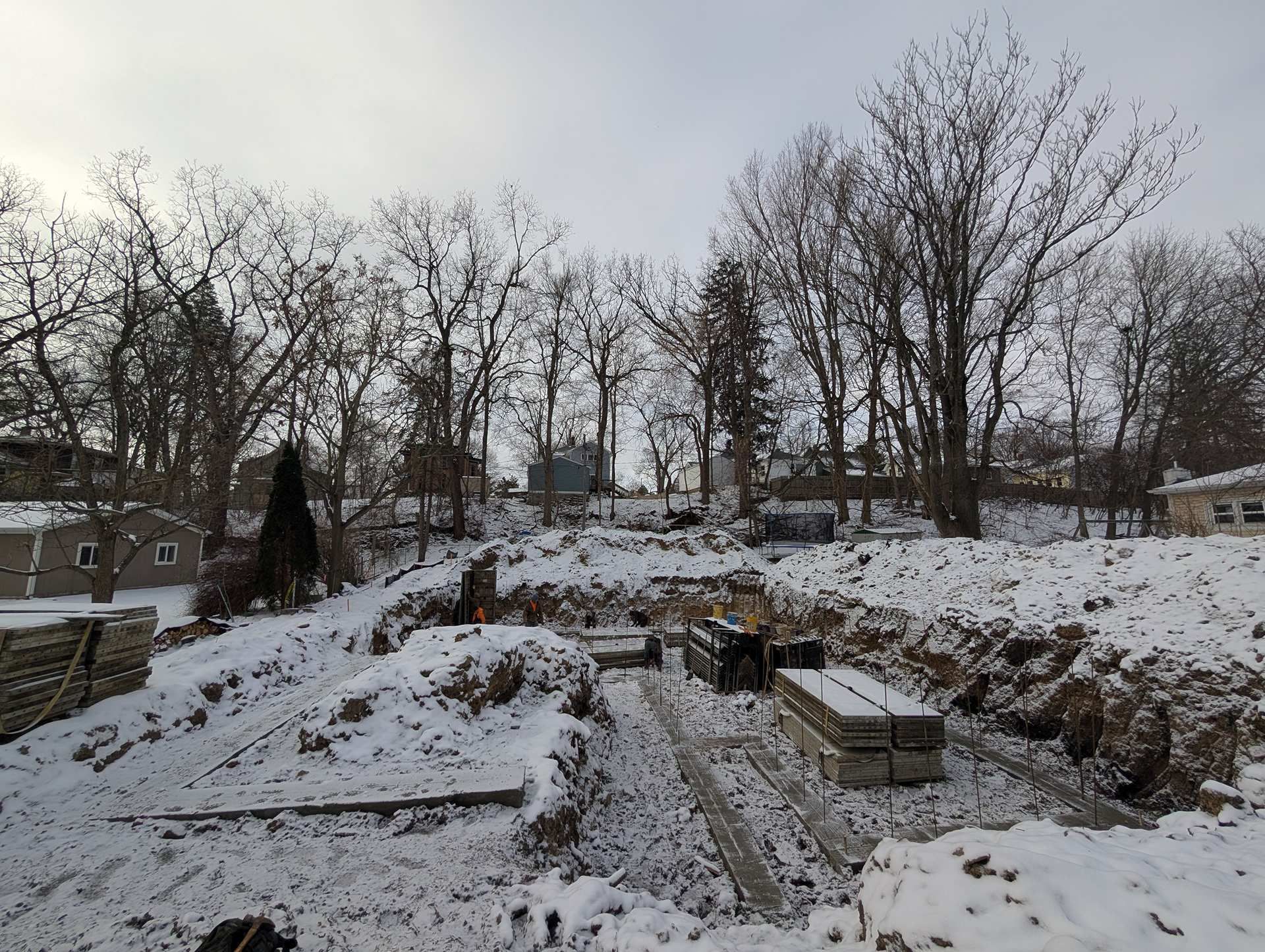 A construction site in winter with a snow-covered excavated trench, foundation materials, and trees in the background.