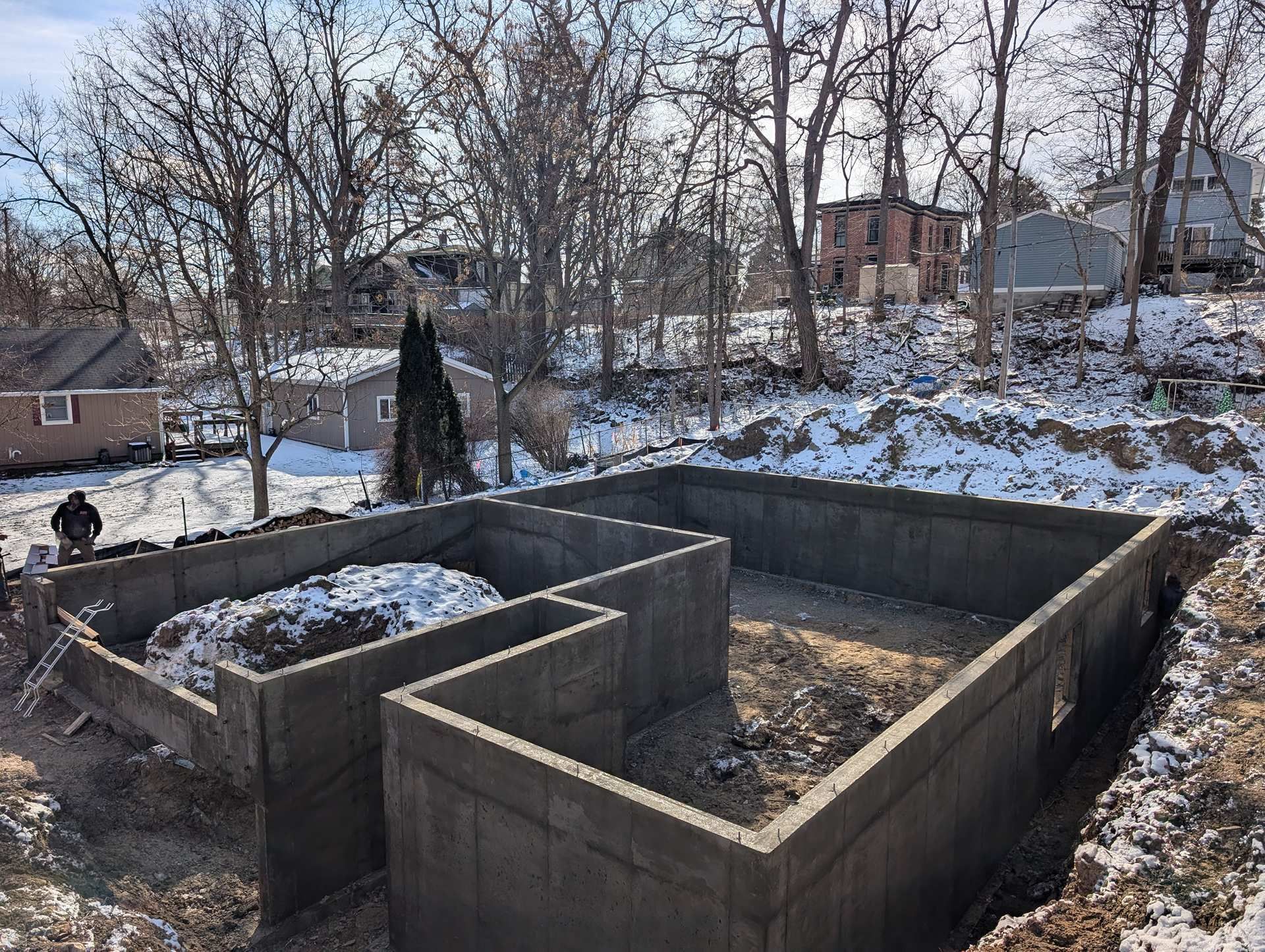 A concrete foundation of a house under construction on a snowy, wooded lot.