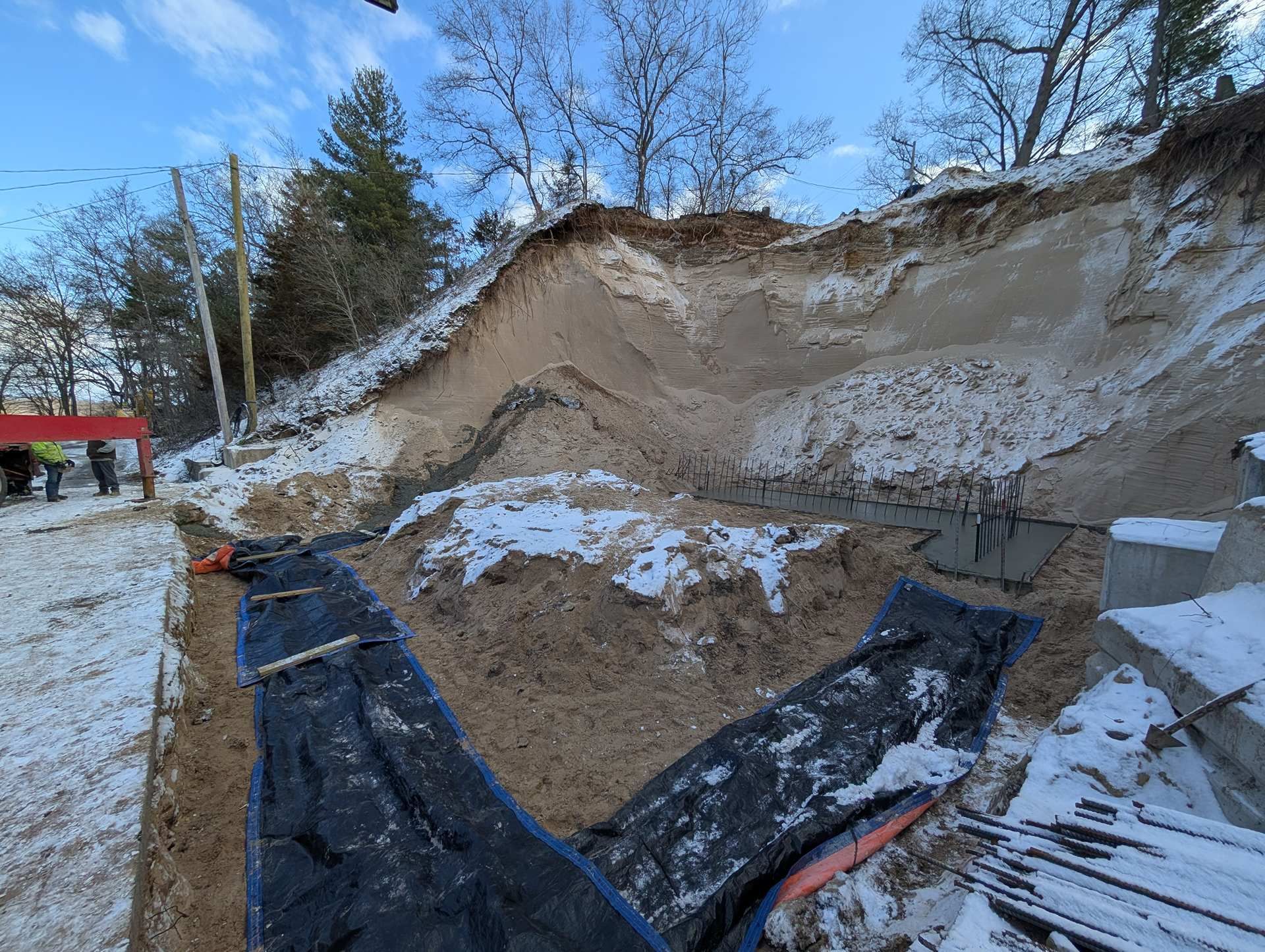 Construction site with a deep excavated pit, concrete footings, and black protective plastic liners, covered in snow.