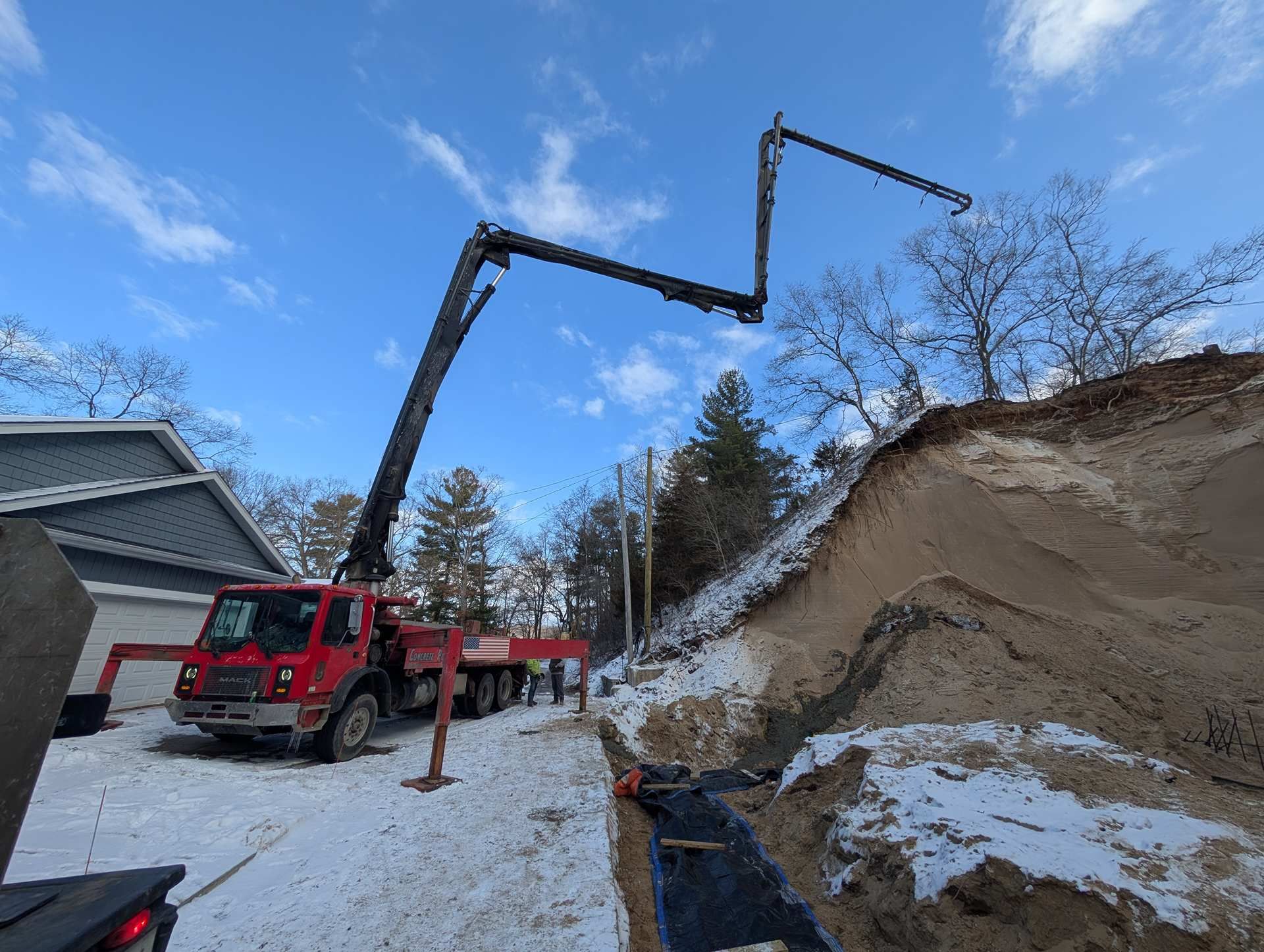 A red concrete pump truck with an extended boom reaches over a snowy construction site near a residential home.