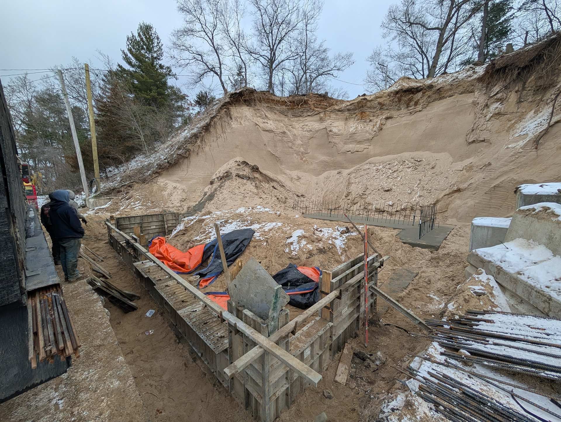 A construction site with foundation forms, rebar, and a large excavated dirt bank under a cloudy, winter sky.