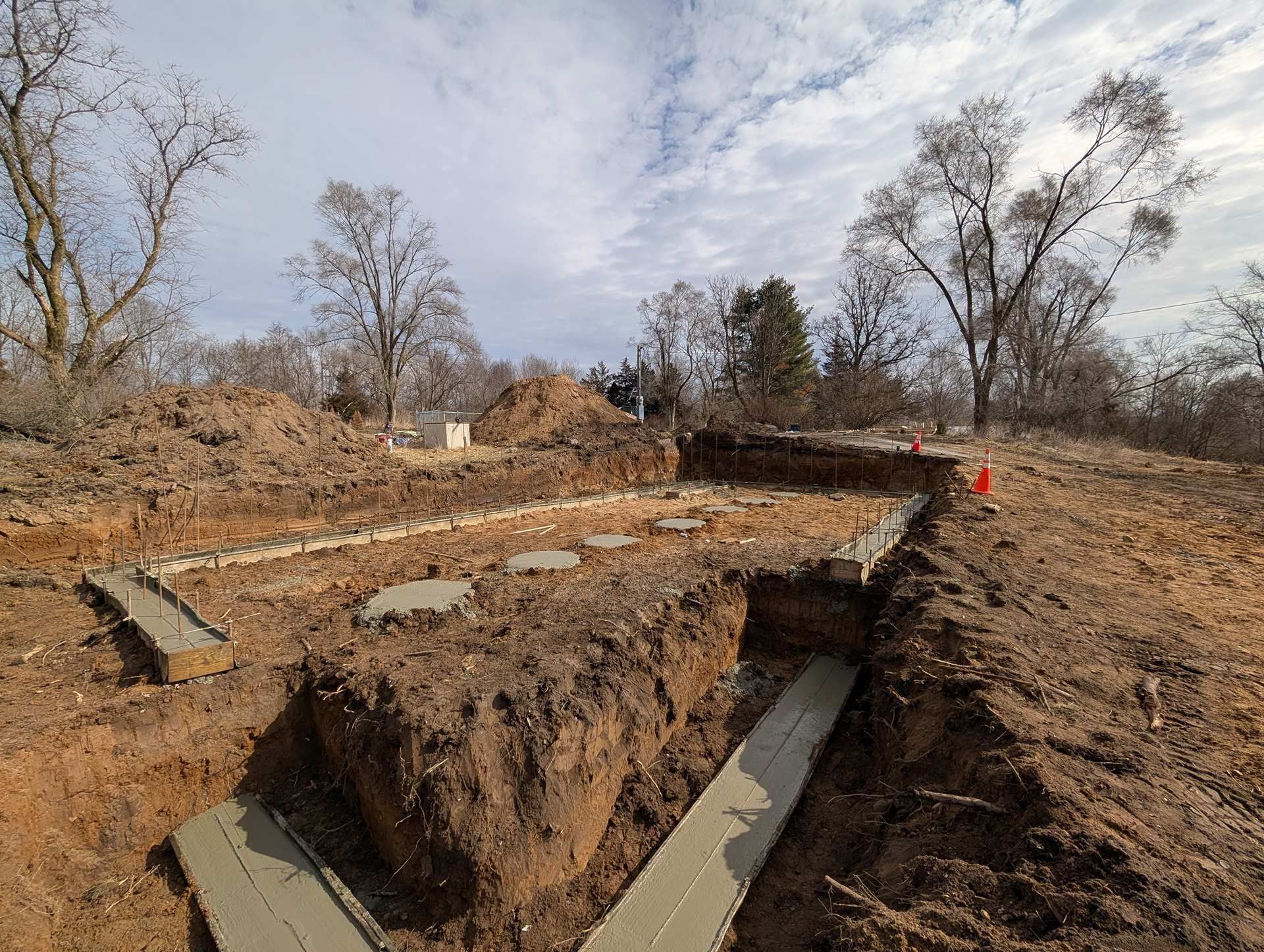 A construction site showing concrete footings poured into dirt trenches at a work area with trees under a cloudy sky.