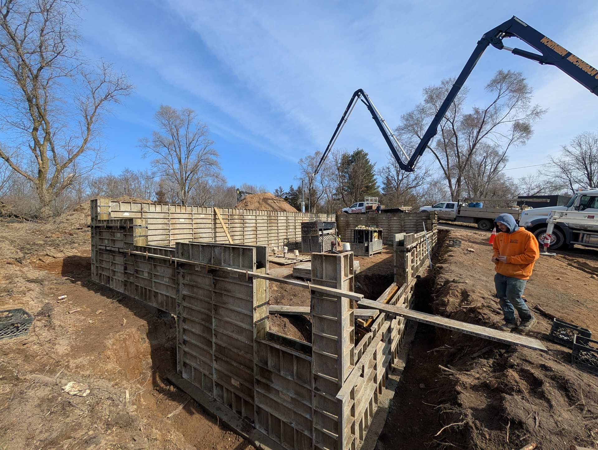 A construction worker in an orange jacket stands near foundation forms while a concrete pump boom extends overhead.