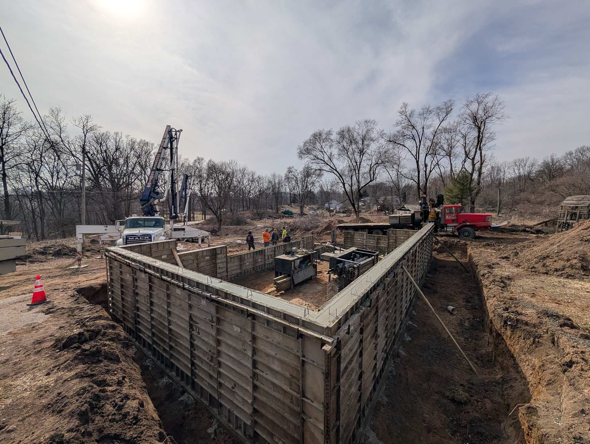 A construction site showing concrete foundation forms for a building, with heavy machinery and bare trees in the background.