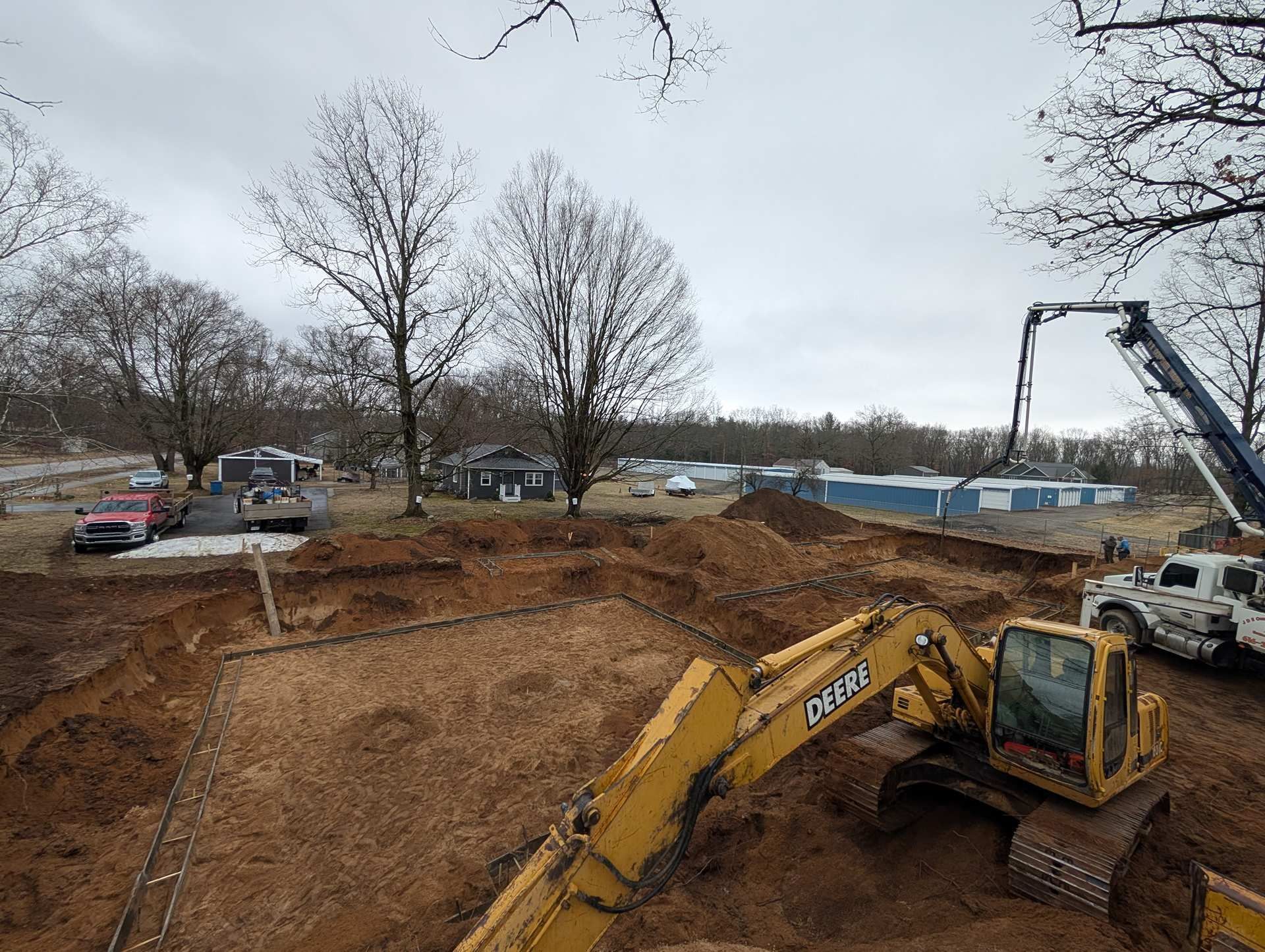 A yellow excavator sits at a construction site with a concrete pump truck, preparing for a building foundation.