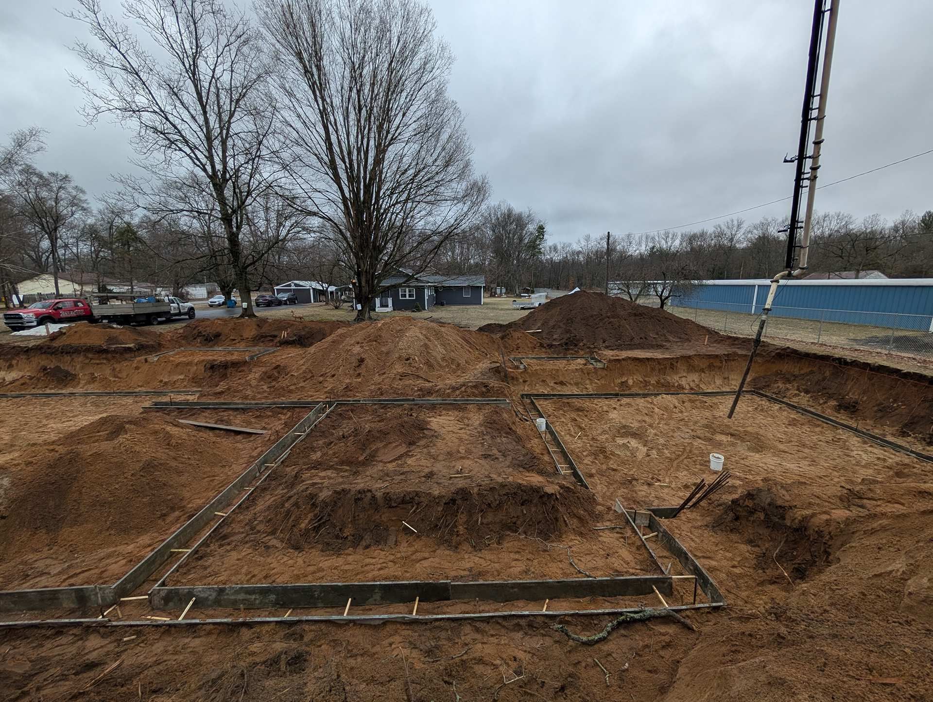 Construction site with wooden footing forms laid out on bare dirt, under an overcast sky.