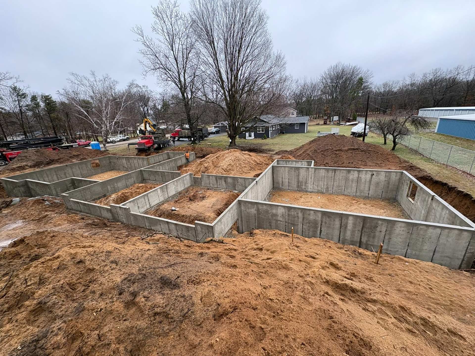 A construction site shows a newly poured concrete foundation for a building, surrounded by piles of dirt on a cloudy day.