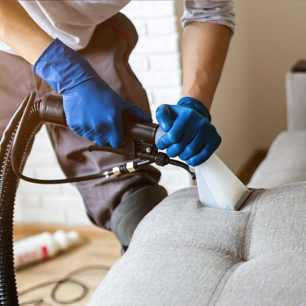 A man wearing blue gloves is cleaning a couch with a vacuum cleaner.