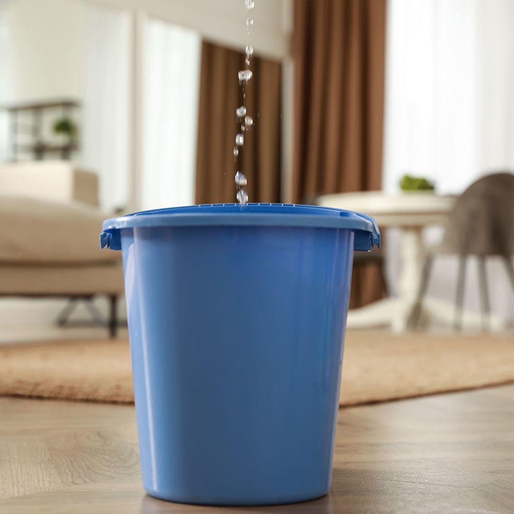 A blue bucket with water pouring out of it in a living room.
