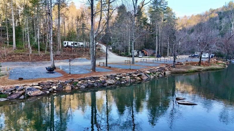 Calm lakeside park with trees, rocky shore, and cabins reflected in the water at dusk