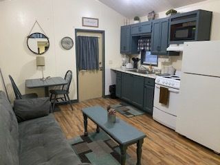 Small living room with gray sofa, glass coffee table, kitchen cabinets, dining table, and white refrigerator.