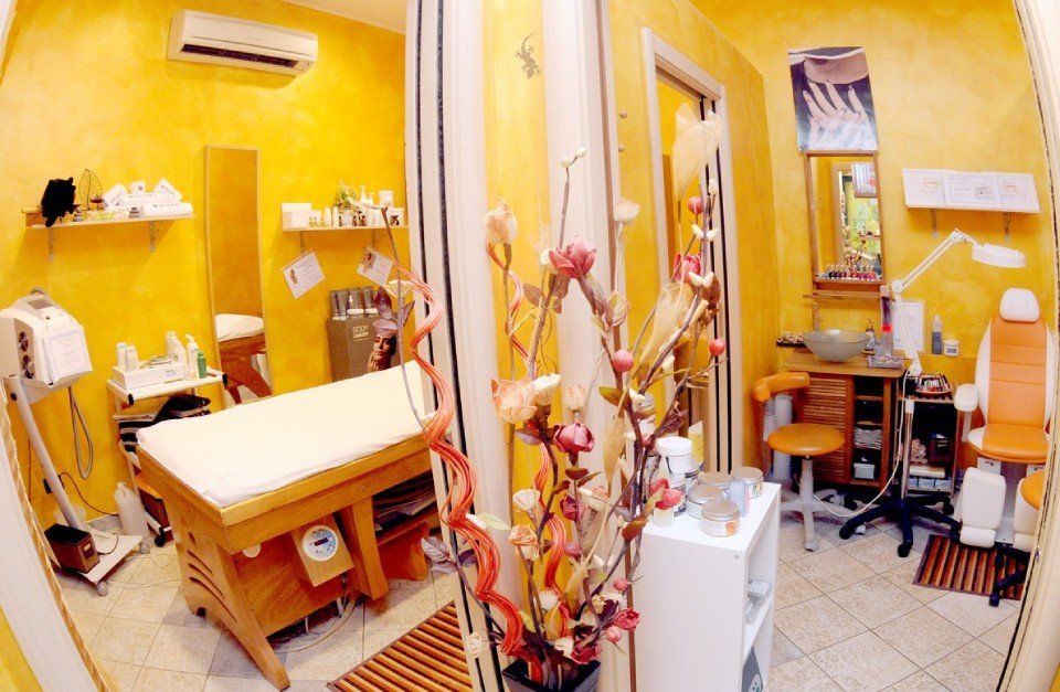 Interno del salone con pareti gialle. Lettino per massaggi e postazione pedicure visibili, fiori sul pavimento.