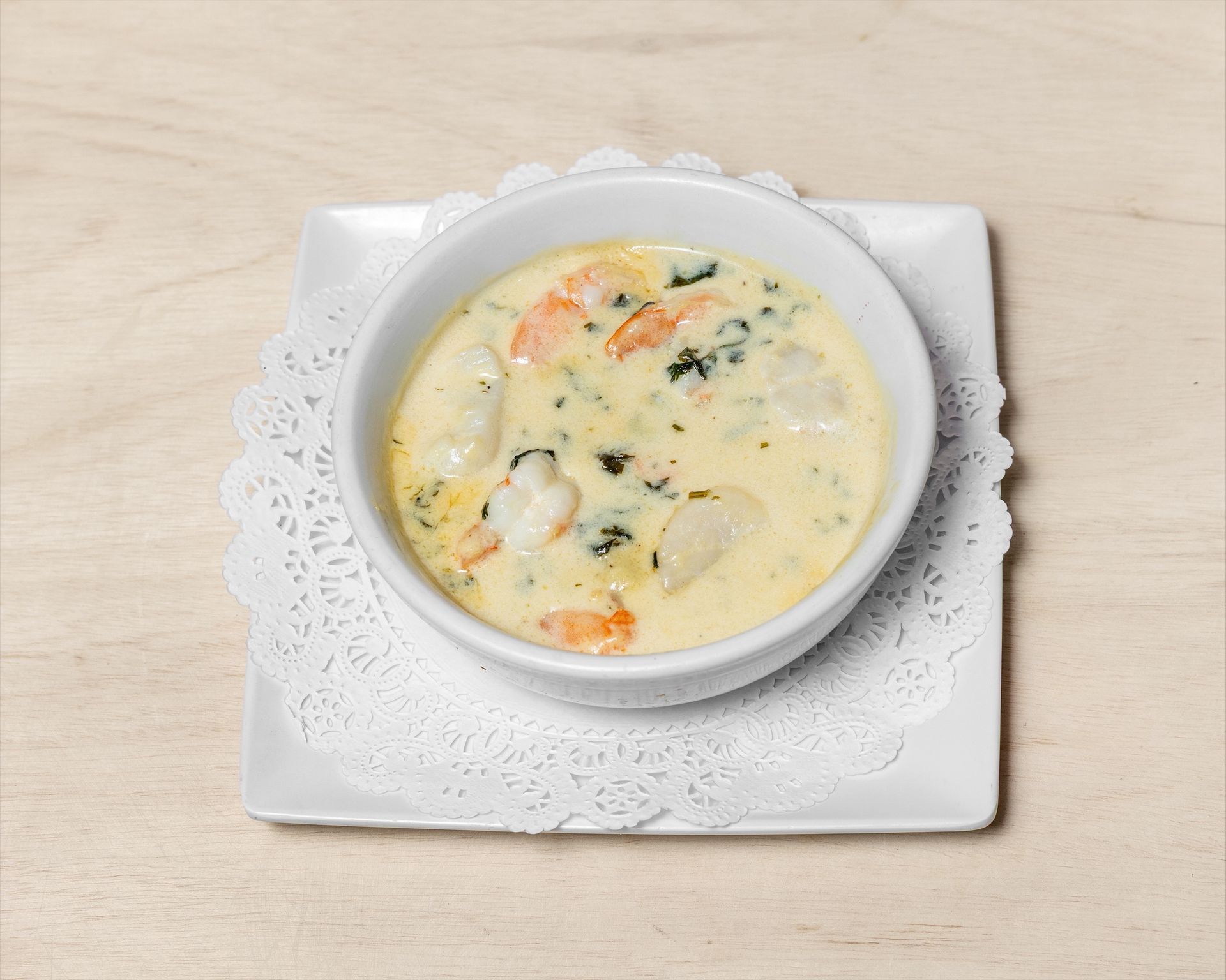Seafood chowder in a white bowl, on a doily and square plate, light wood background.