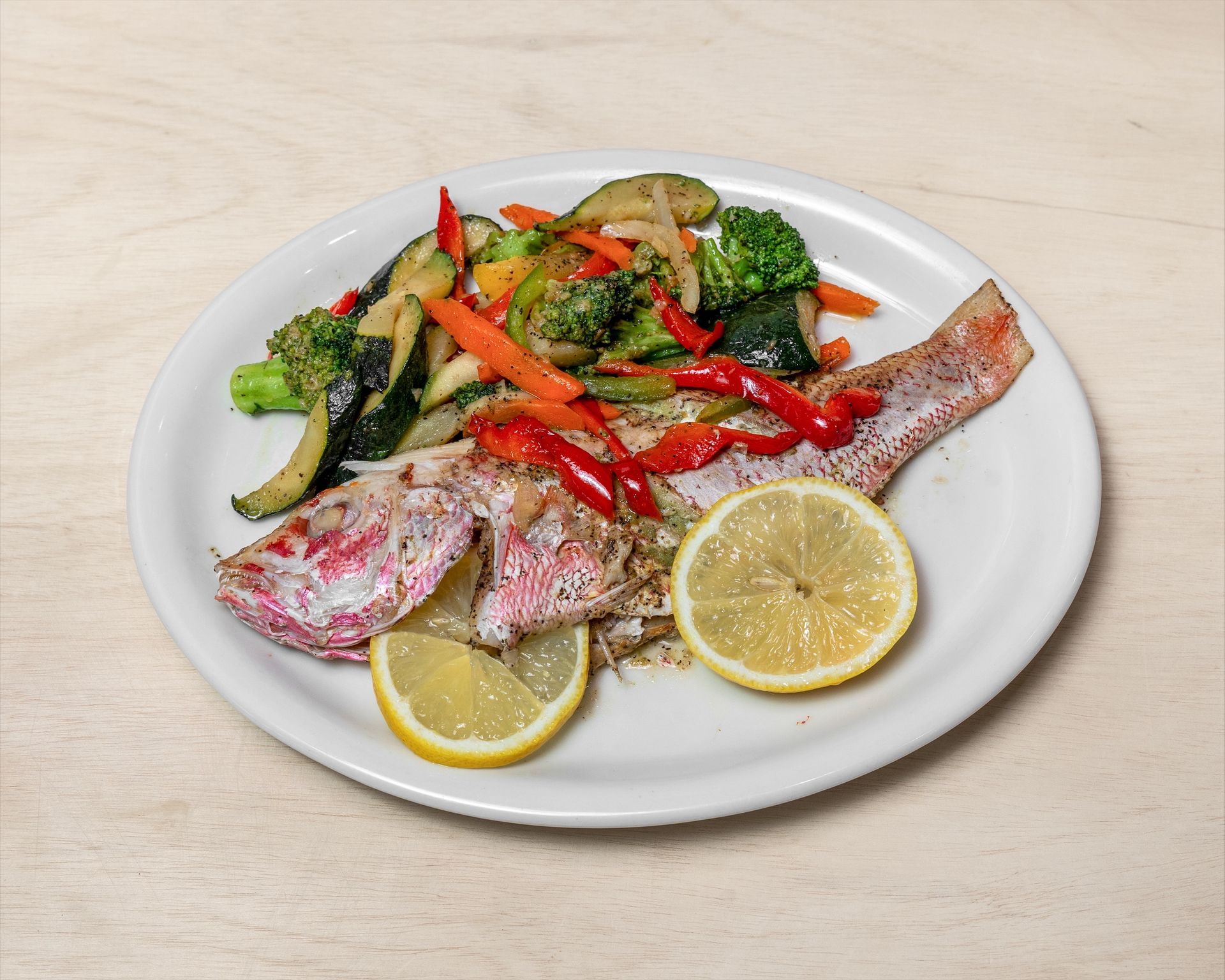 Grilled fish with vegetables and lemon slices on a white plate.
