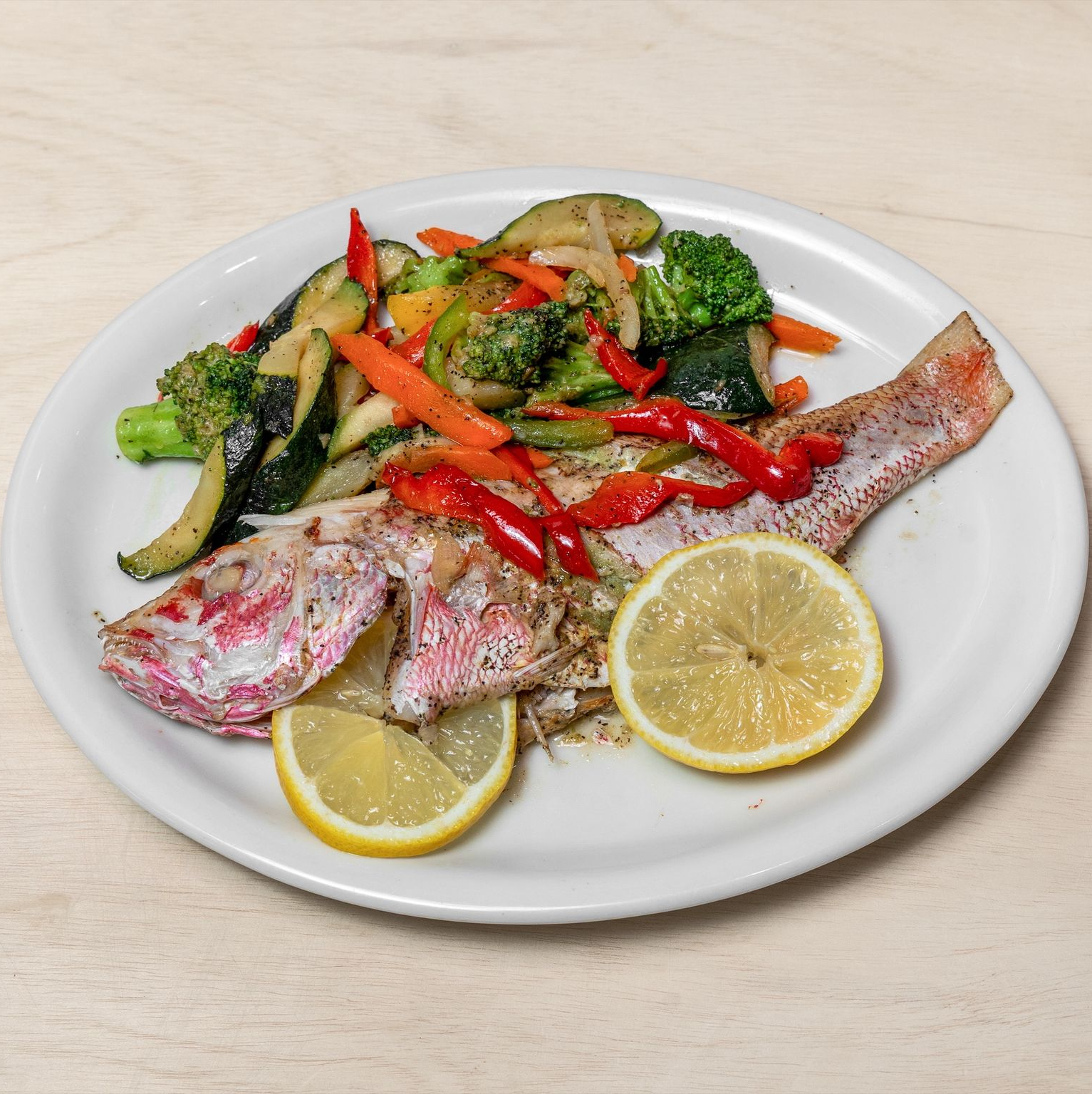 Grilled red snapper with vegetables and lemon slices on a white plate.