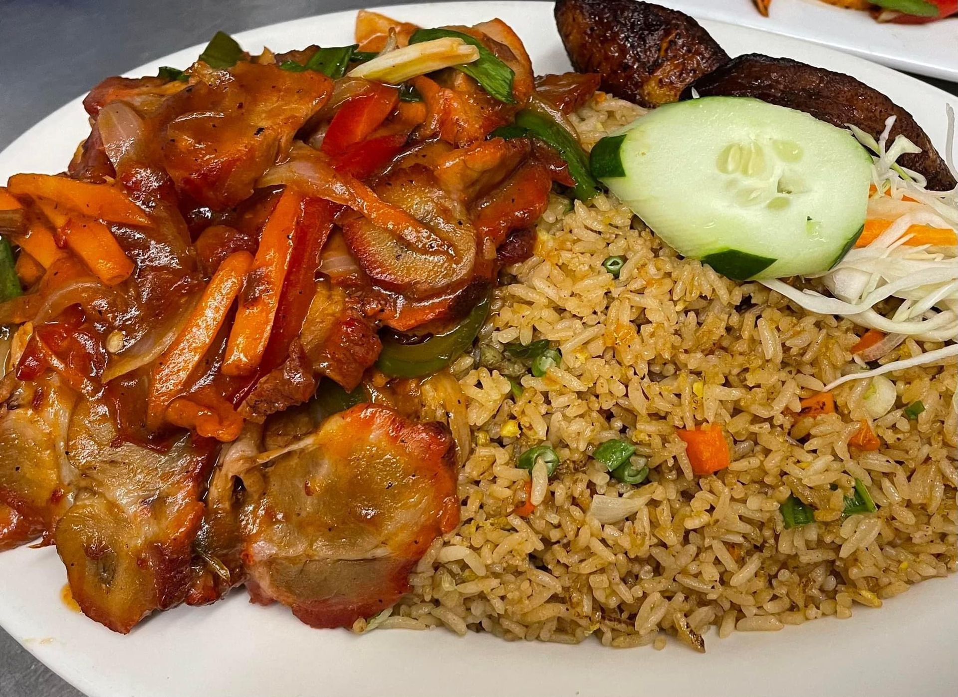 Plate of fried rice, sauced pork with vegetables, plantains, cucumber, and slaw.