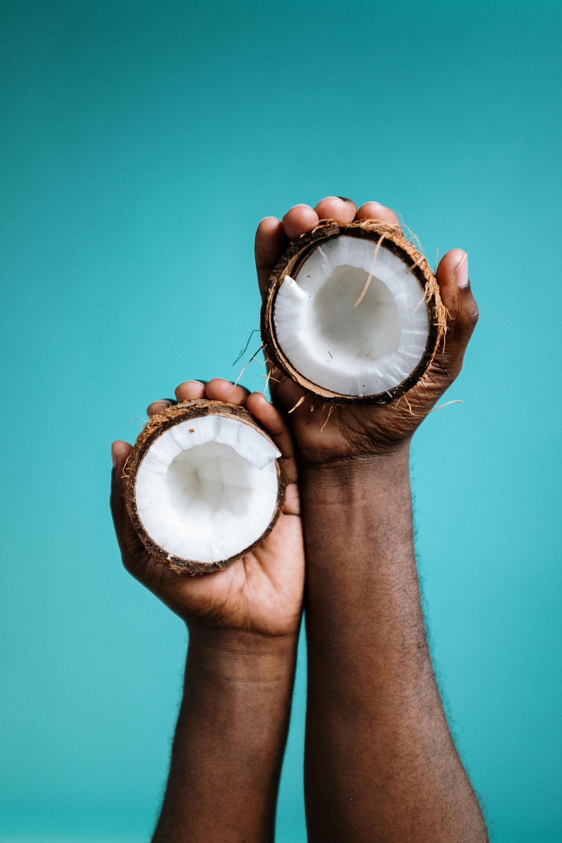 Hands holding halves of a coconut, showcasing white flesh against a teal background.