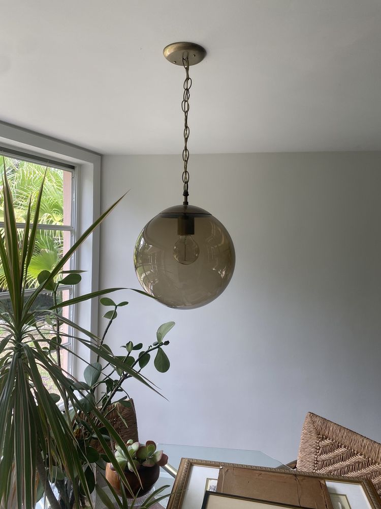 Round, smoky glass pendant light hanging from a chain; plants and window visible in the background.