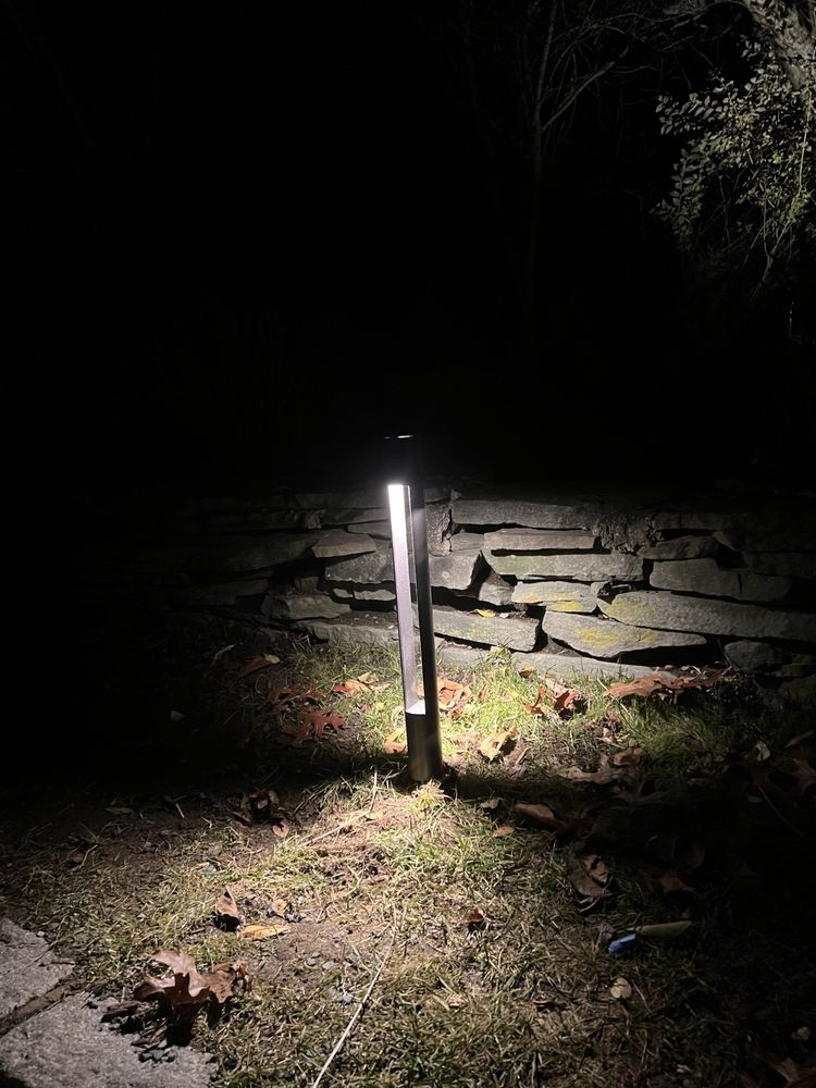 A lit outdoor landscape light illuminates the ground in front of a stone wall at night.