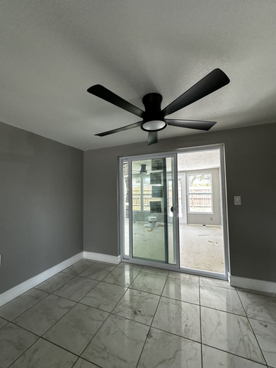 Empty room with a gray wall, sliding glass door, and black ceiling fan. Marble-look floor.