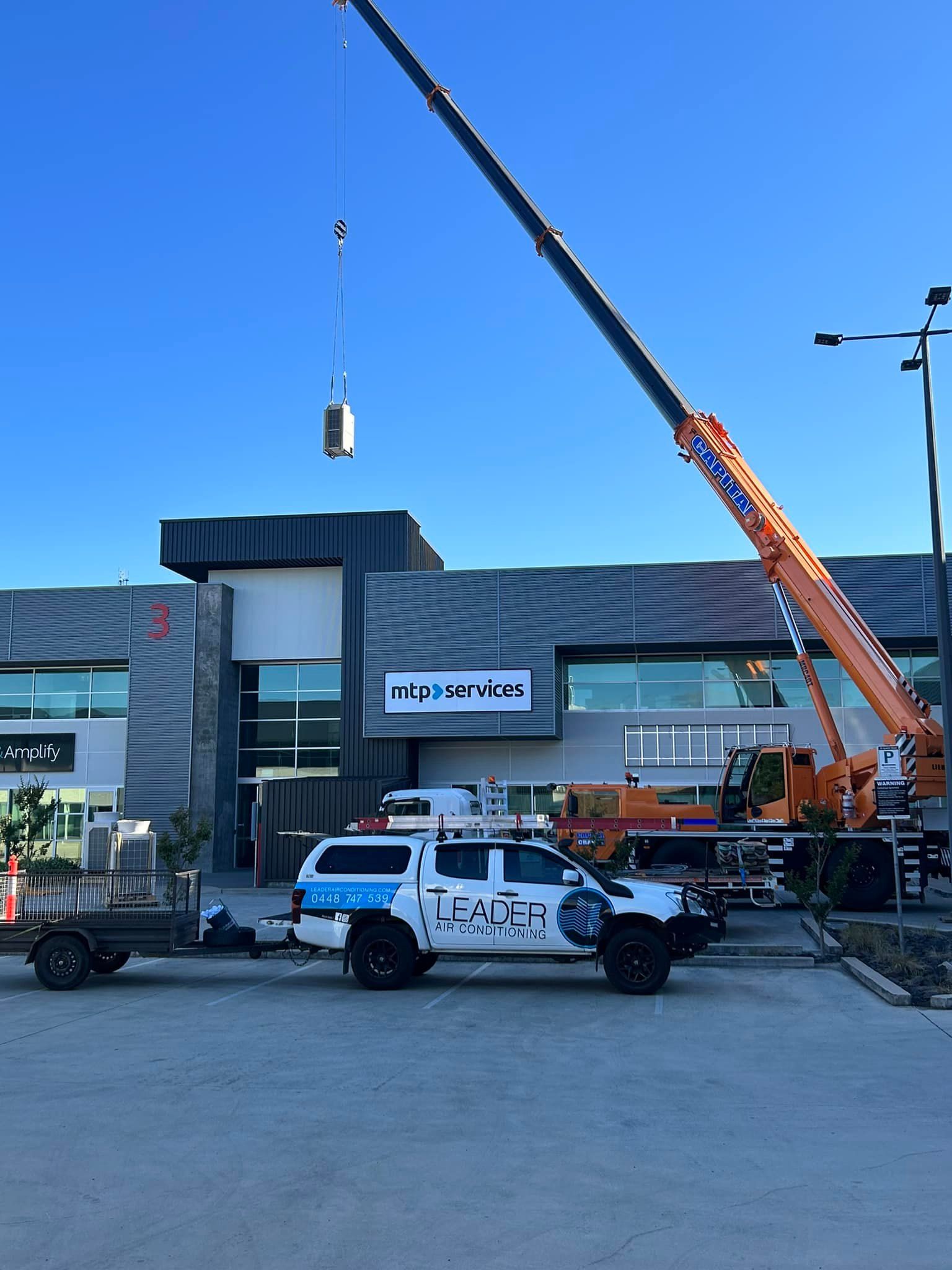 Crane lifting a container near a white truck with blue decals and a modern building under a clear blue sky. — Leader Air Conditioning in Queanbeyan, NSW
