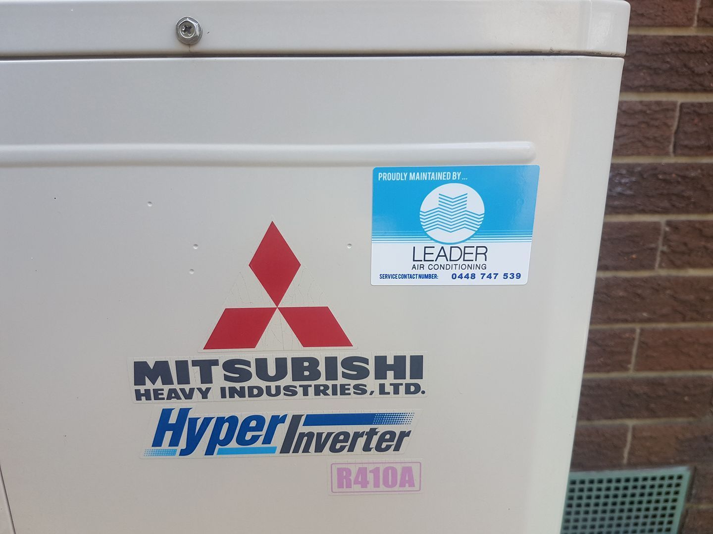 Mitsubishi air conditioning unit with logos and stickers, mounted on an exterior wall. — Leader Air Conditioning in Queanbeyan, NSW